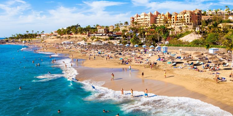 Foto de la playa de Adeje donde se puede ver la calidad de la arena, el color del mar azul turquesa y al fondo todos los alojamientos entre los que se encuentran algunos apartamentos que admiten perros GRATIS
